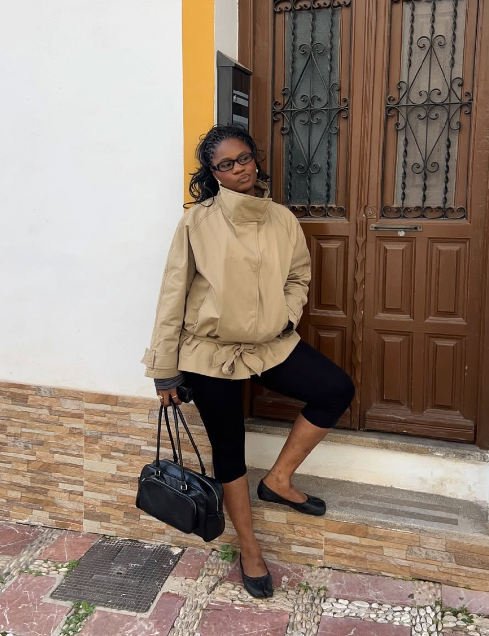 Modelo posa com jaqueta de gola alta bege de caimento amplo, legging preta e bolsa estruturada diante de porta marrom.