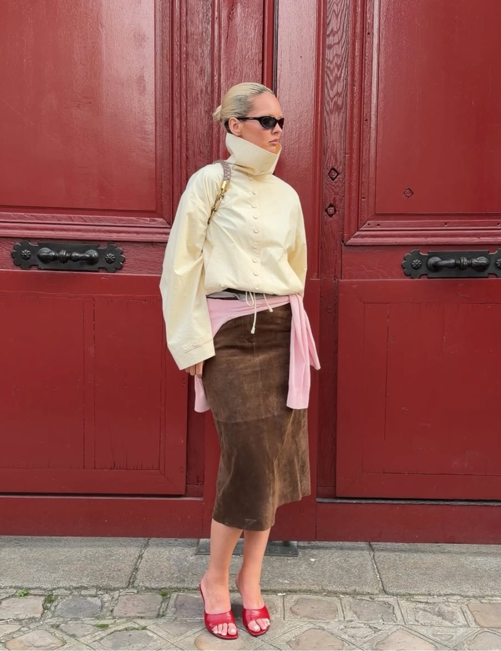 Modelo posa com jaqueta de gola alta creme, saia marrom aveludada e sandálias vermelhas diante de porta vinho.