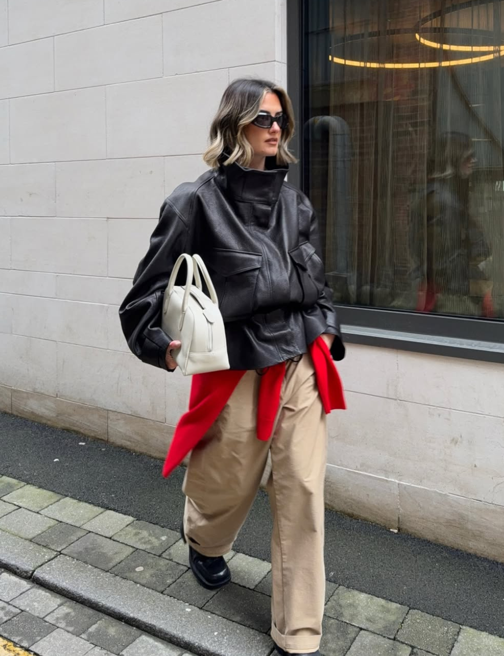 Modelo caminha com jaqueta de gola alta preta em couro, calça bege ampla, bolsa off-white e toque vermelho elegante.