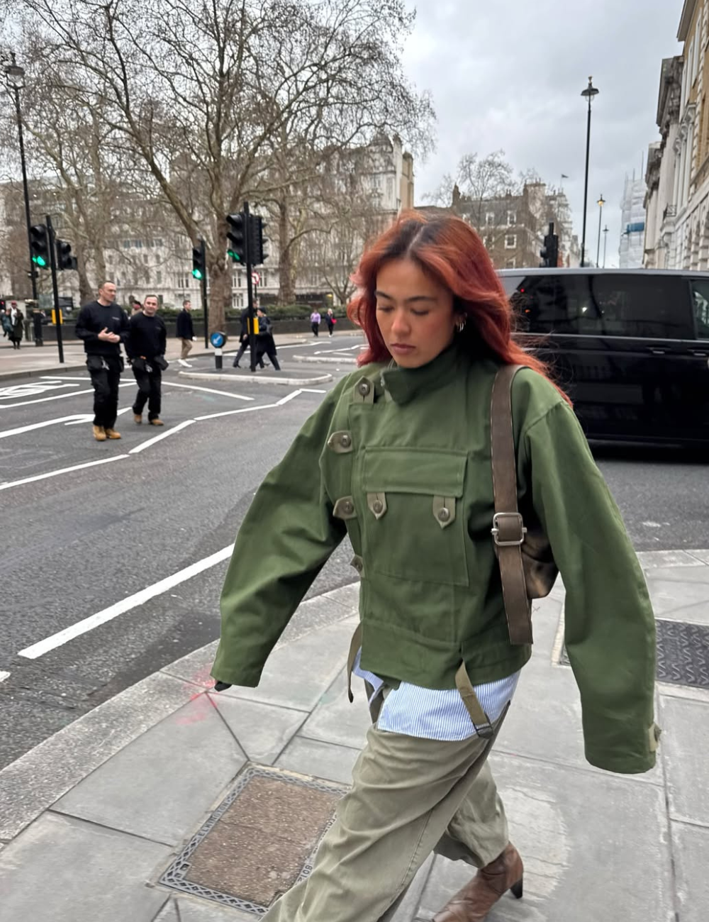 Modelo caminha com jaqueta de gola alta verde utilitária, calça ampla bege e bolsa marrom em rua urbana cinza.
