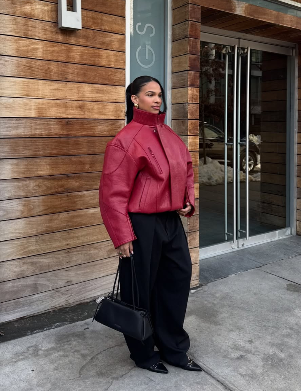 Modelo posa com jaqueta de gola alta vinho em couro, calça preta ampla e bolsa estruturada diante de fachada de madeira.
