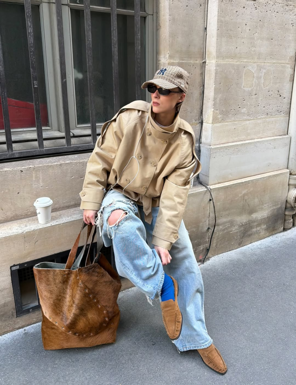 Modelo posa com jaqueta de gola alta bege, jeans rasgado e boné xadrez diante de parede urbana, em clima casual cool.