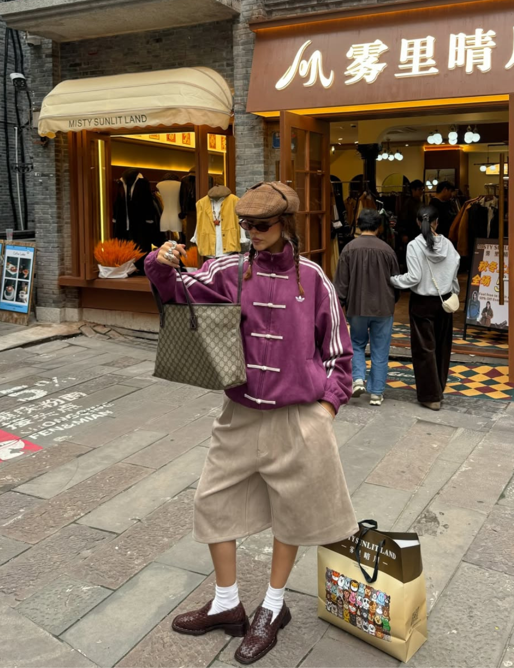 Looks com jaqueta nó chines roxa, modelo posa com boina e bermuda bege diante da loja.