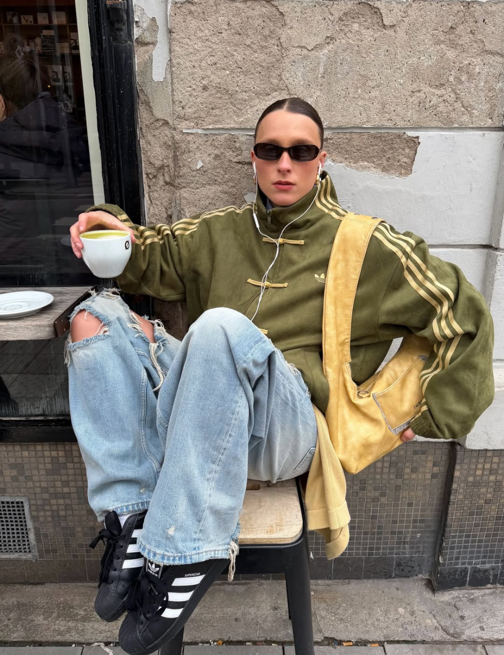 Looks com jaqueta nó chines verde oversized, jeans rasgado e bolsa amarela criam mood urbano despojado; modelo posa sentada.