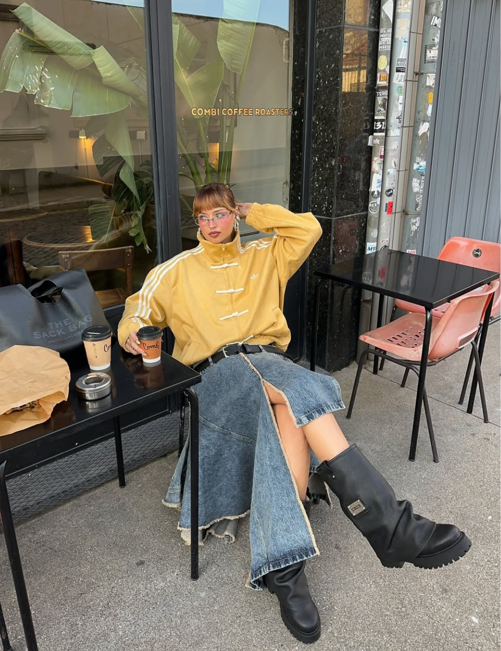 Looks com jaqueta nó chines: modelo posa sentada com jaqueta amarela, saia jeans longa e botas pretas em café urbano.