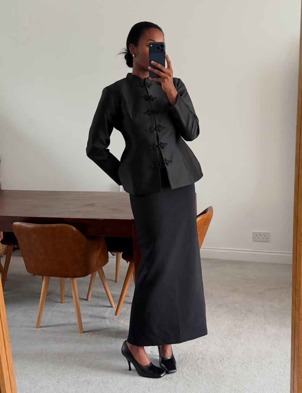 Looks com jaqueta nó chines: modelo posa de preto com jaqueta acinturada, saia longa e salto em ambiente minimalista.