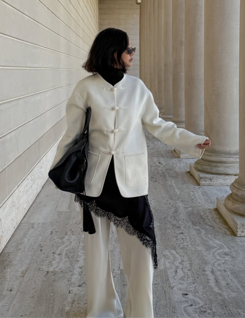 Looks com jaqueta nó chines: modelo posa de perfil com casaco branco, calça ampla, renda preta e bolsa preta em colunata.