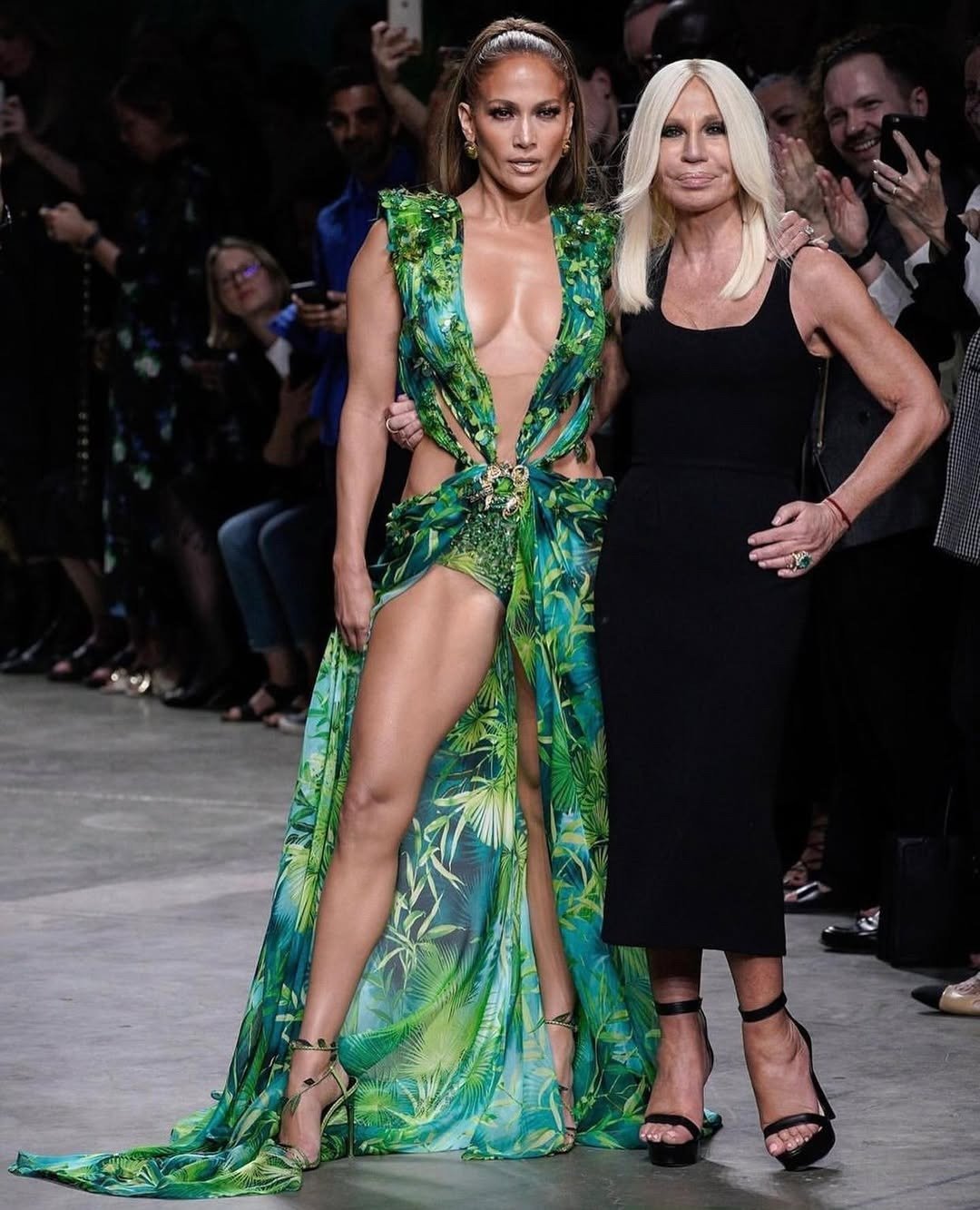 Modelo posa com vestido Versace verde tropical de tecido fluido, decote profundo e fenda alta, ao lado de mulher de vestido