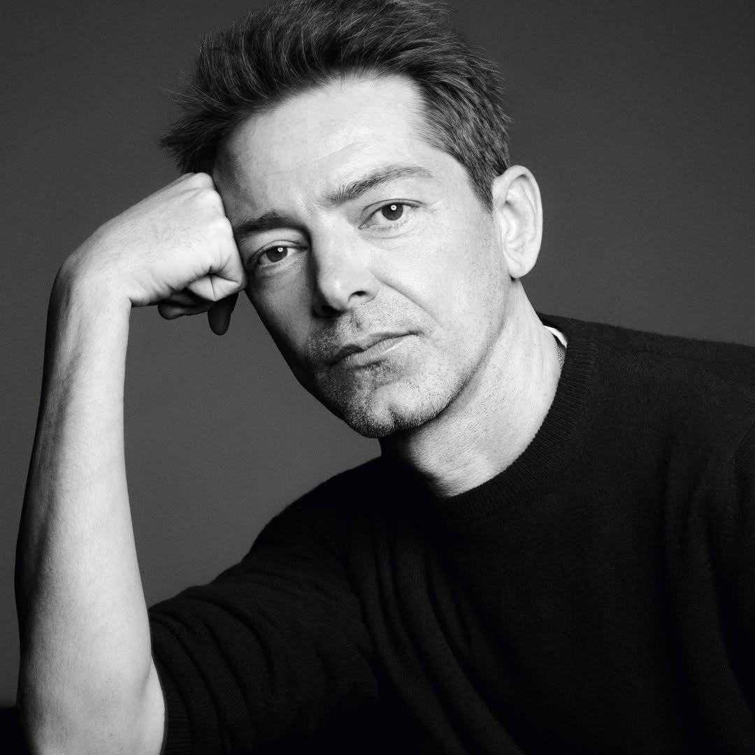 Homem posa pensativo com suéter preto de malha lisa, expressando sobriedade e elegância minimalista no estilo Versace.