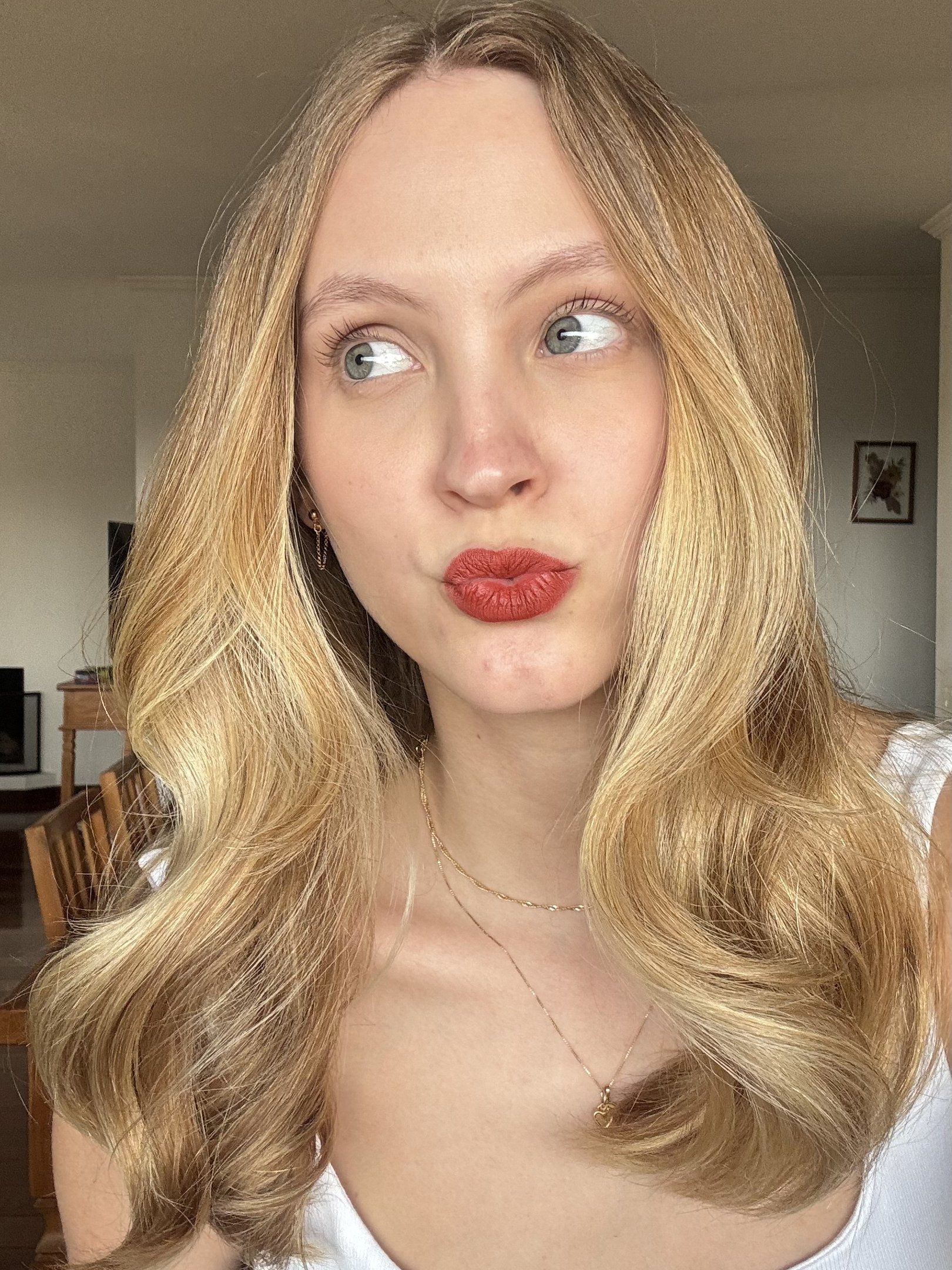 Modelo posa com lápis labial da Nars terracota, regata branca e ondas loiras em clima leve e elegante.