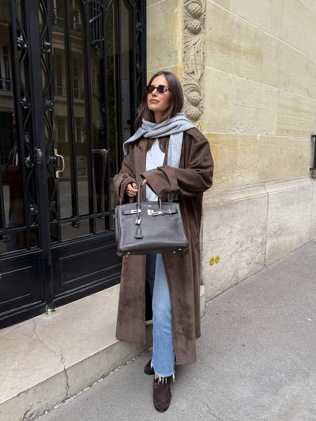 Modelo posa com mocassim feminino com calça jeans clara, trench marrom e malha cinza em clima urbano elegante.