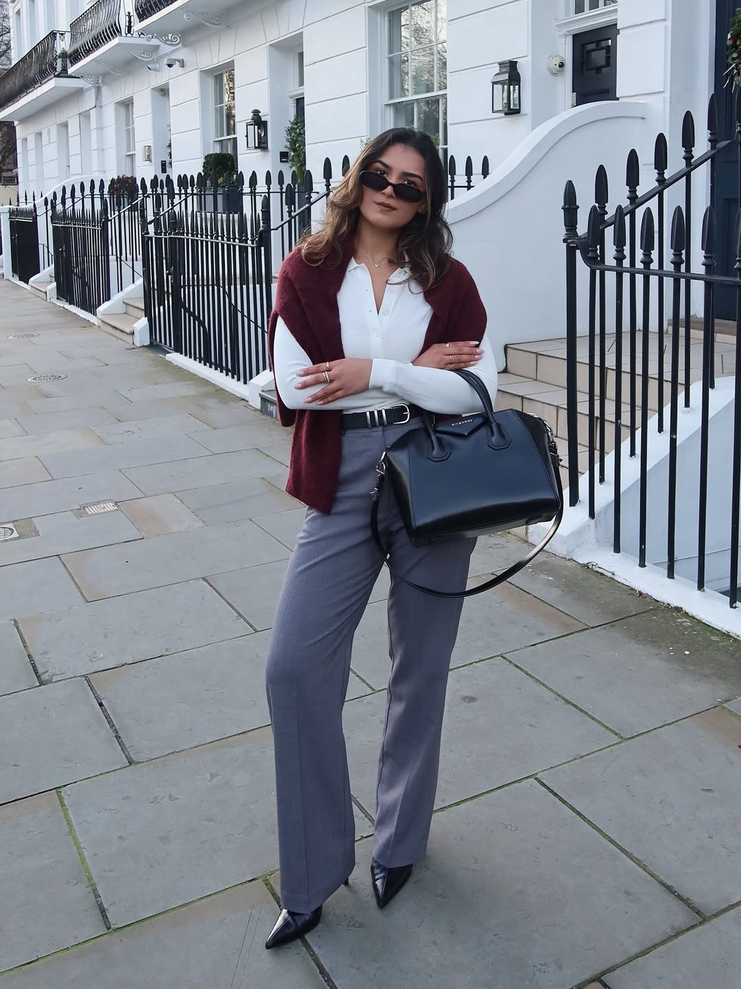 Mulher elegante com tricô vinho, blusa branca, calça cinza e bolsa preta em rua clássica. Roupas de armário capsula feminino.