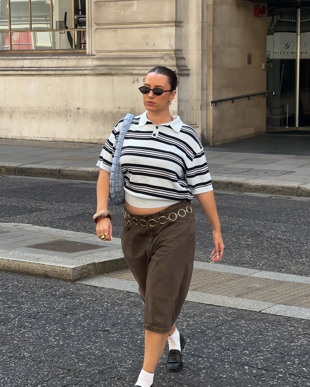 Look de festival urbano: modelo caminha com polo listrada cropped, bermuda marrom ampla e corrente, clima cool.