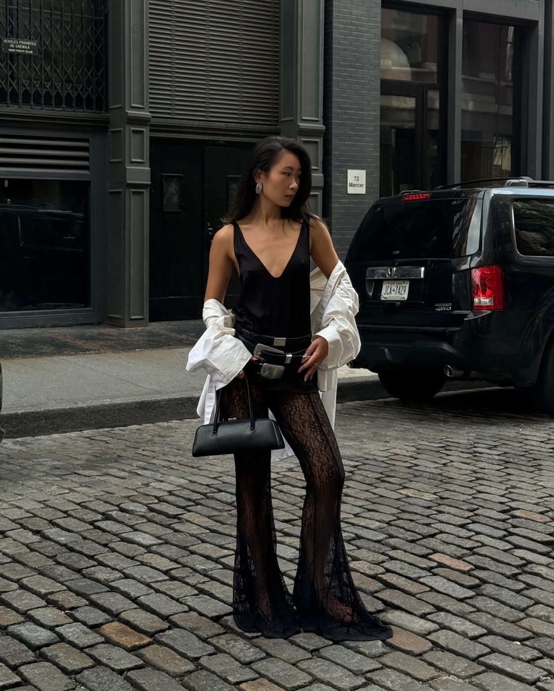 Look de festival com body preto, calça rendada, camisa branca solta e bolsa preta; modelo posa com atitude urbana.