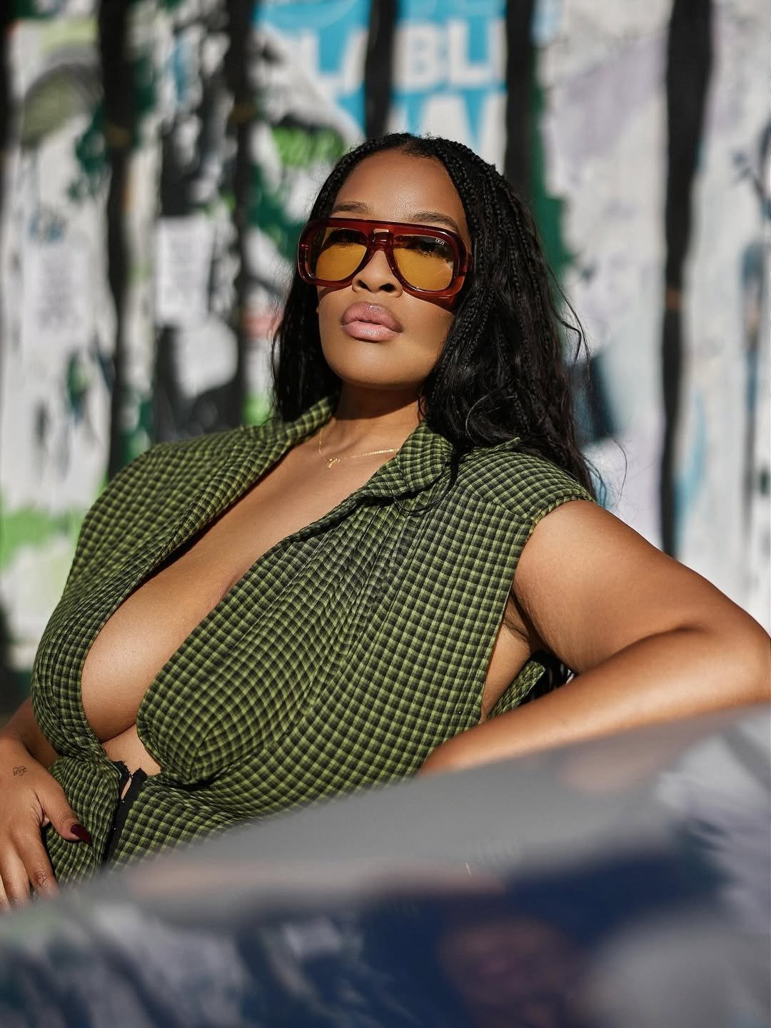 Look de festival: modelo posa com vestido xadrez verde decotado e óculos âmbar oversized diante de muro grafitado.