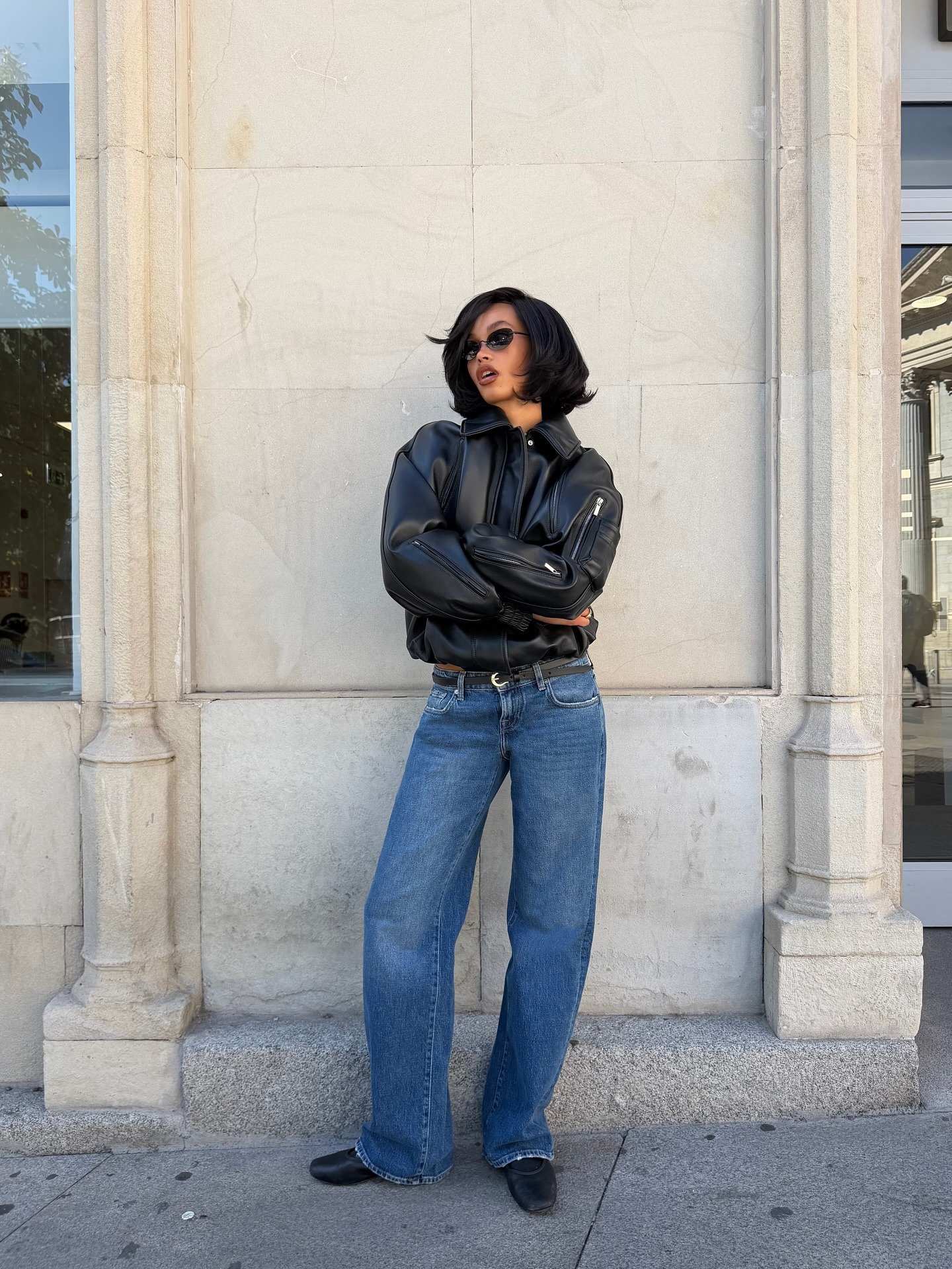 Look minimalista com modelo posa de braços cruzados em jaqueta preta oversized e jeans azul amplo diante de parede de pedra.