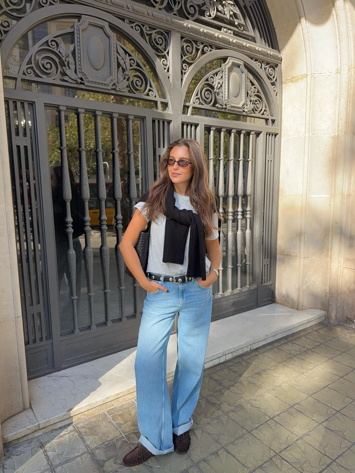 Look minimalista com modelo posa de jeans amplo, camiseta cinza e tricô preto sobre os ombros em clima elegante.