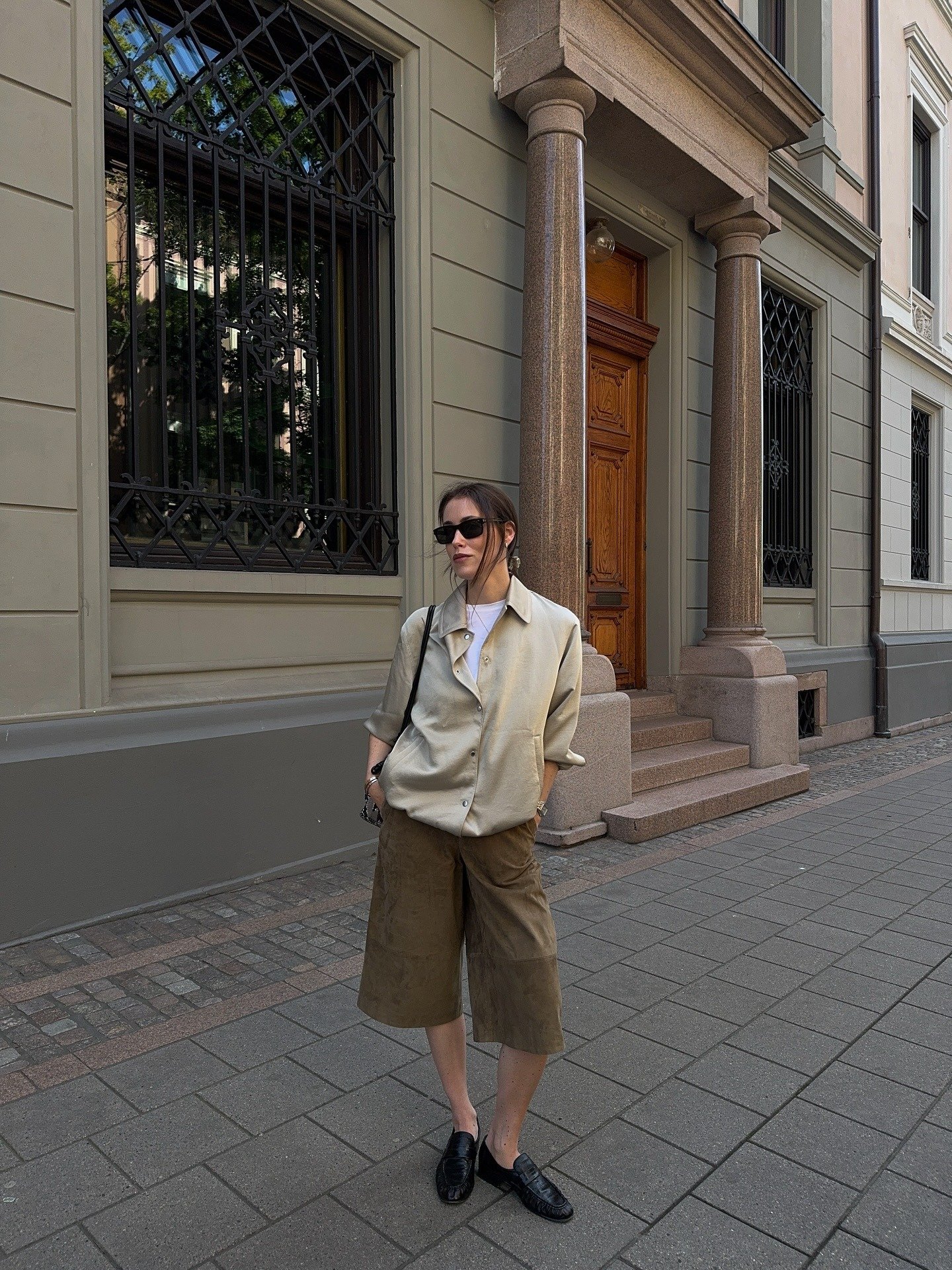Look minimalista com modelo posando de camisa bege ampla, bermuda marrom e mocassins pretos em rua elegante e serena