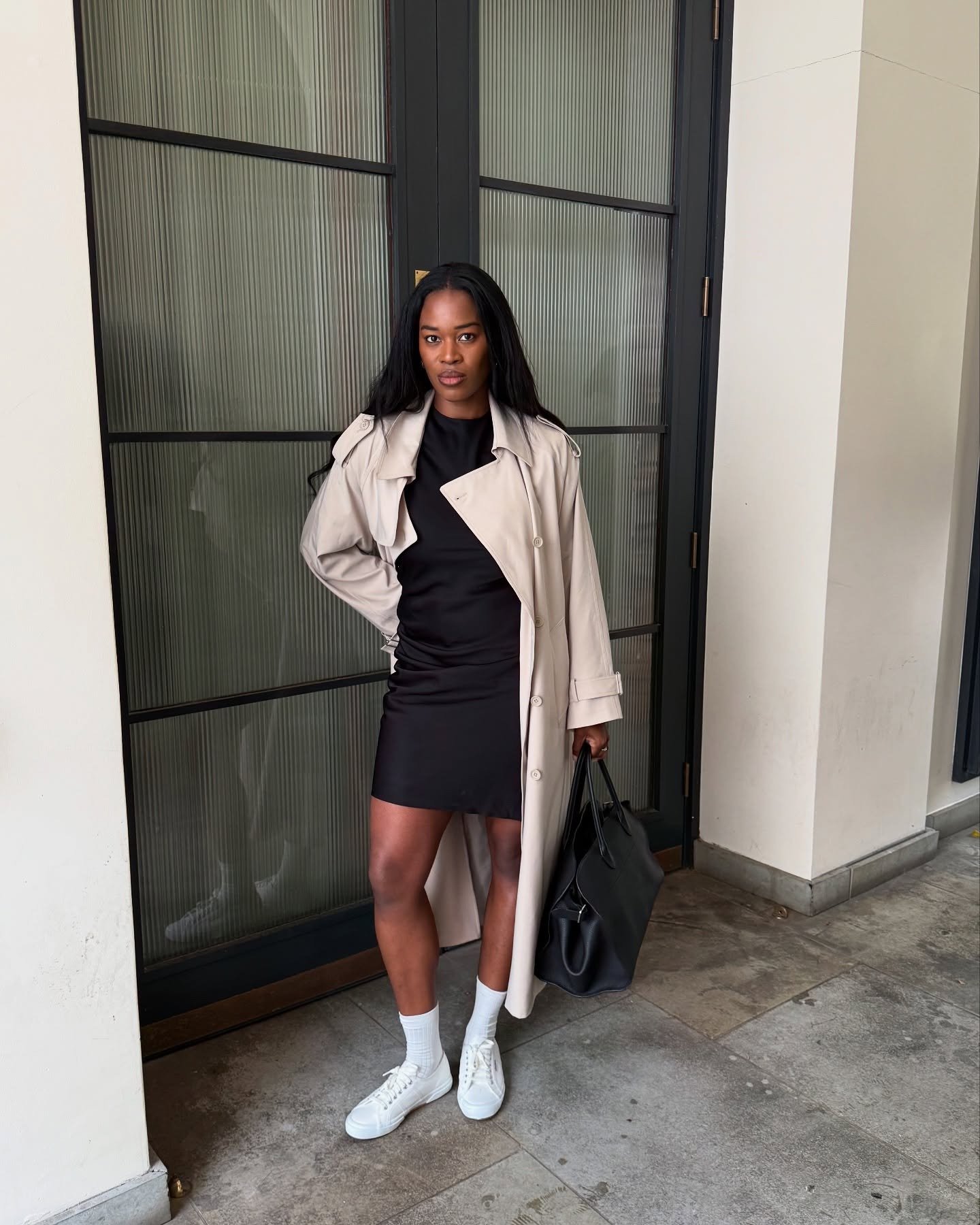 Look minimalista com modelo posando de vestido preto, trench bege e tênis branco diante de porta canelada elegante