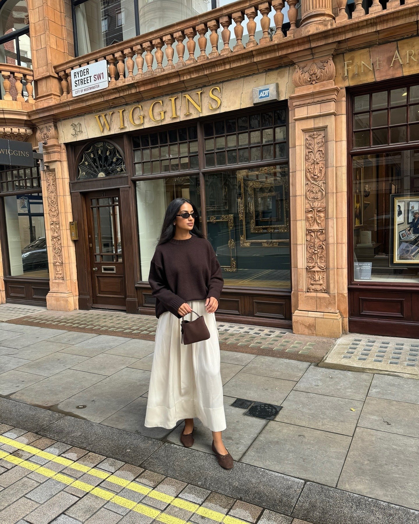 Look minimalista com tricô marrom e saia acetinada off-white; modelo posa serena na rua.