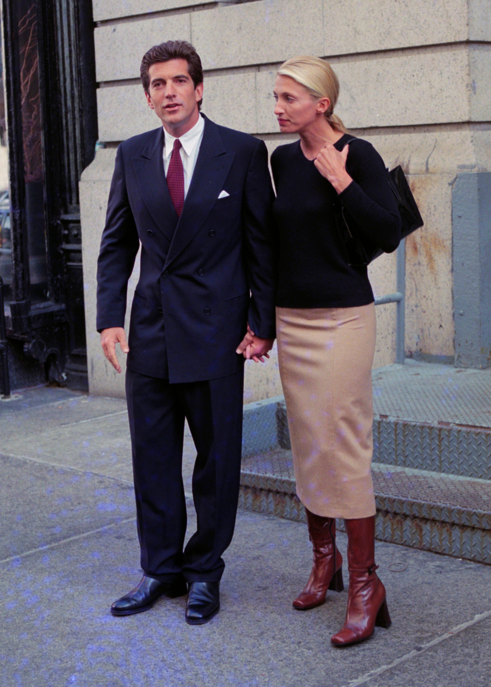 Carolyn Bessette exibe look minimalista com suéter preto justo, saia lápis bege de tecido liso e botas de couro marrom.