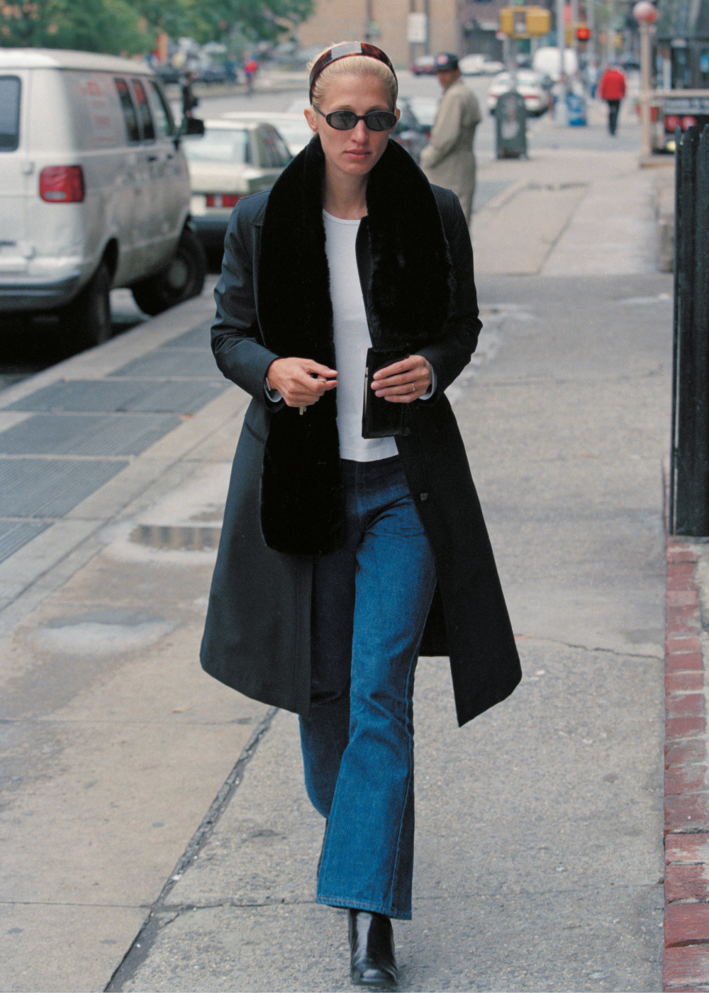Carolyn Bessette exibe looks da Carolyn Bessette com casaco longo preto de gola peluda, jeans flare, blusa branca.