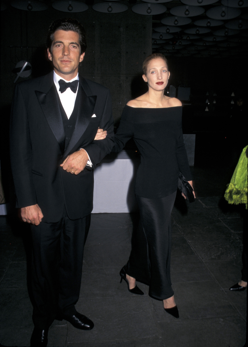 Carolyn Bessette exibe look minimalista com blusa preta ombro a ombro e saia longa de cetim, postura elegante e segura.