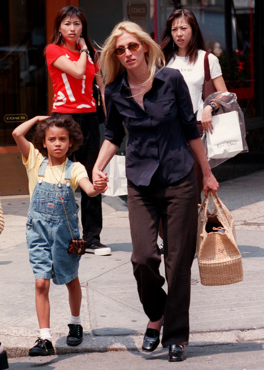 Carolyn Bessette caminha confiante com camisa preta ajustada, calça reta escura e bolsa de palha.