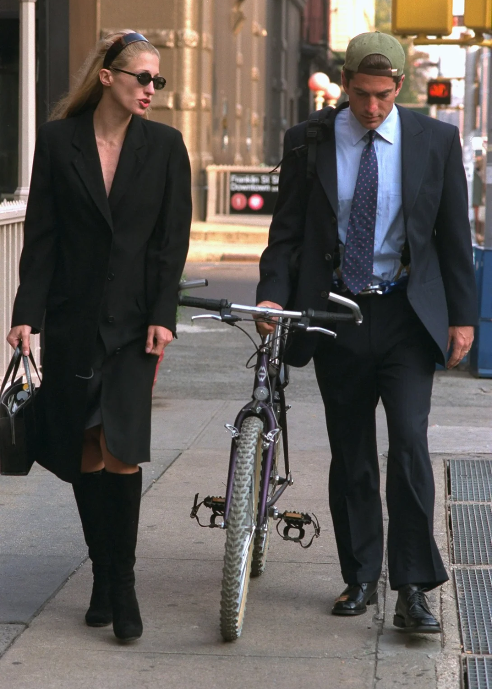 Carolyn Bessette usa sobretudo preto oversized com vestido e bota de cano alto, postura confiante em passeio urbano.