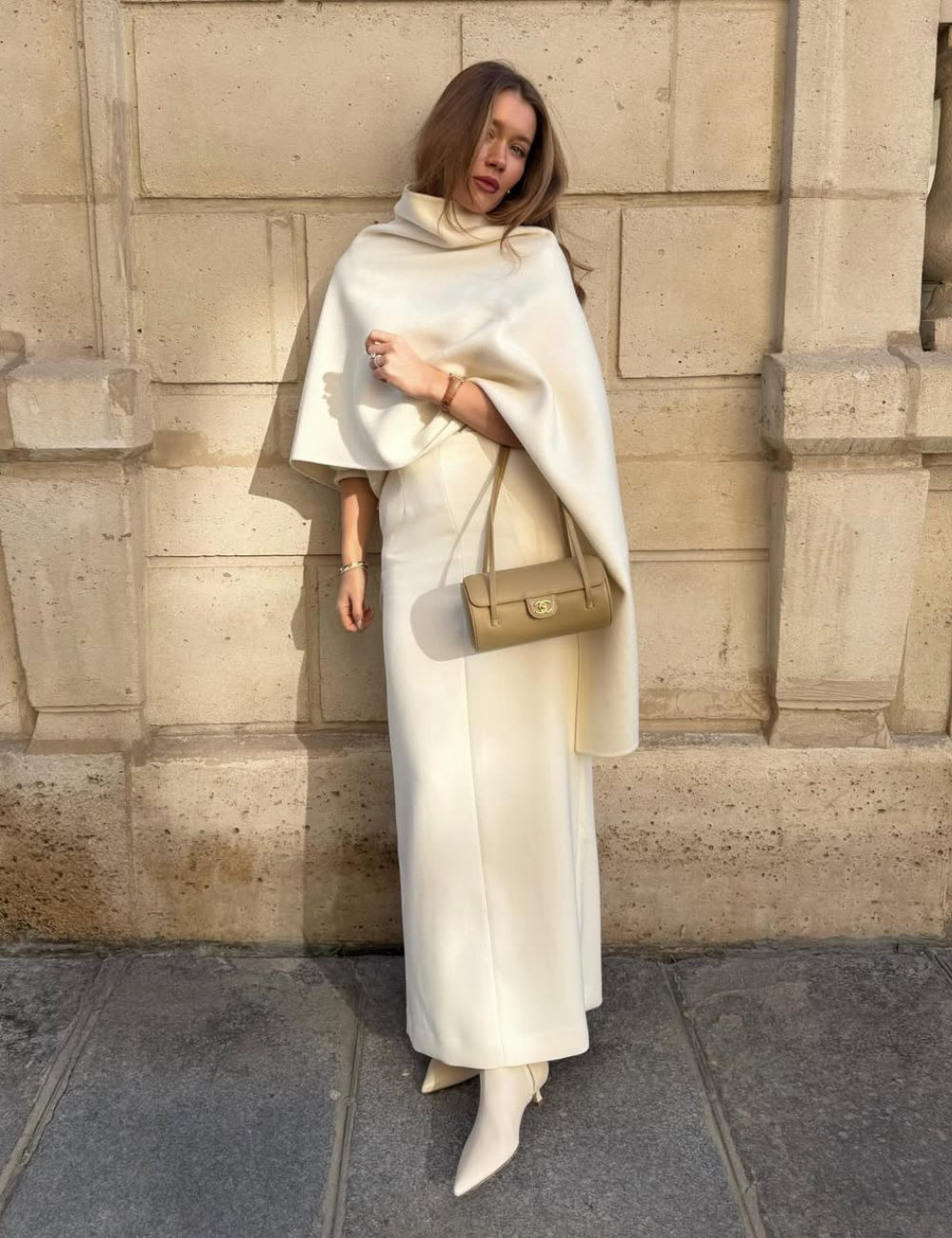 Modelo posa com look monocromático em tons de creme, usando vestido longo e capa de tecido estruturado.