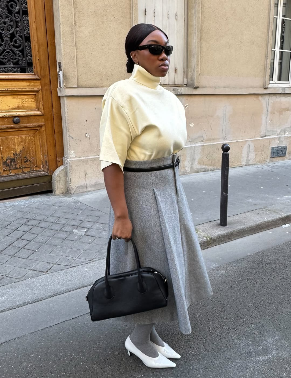 Looks com meia-calça: modelo posa elegante com tricô creme, saia cinza evasê, bolsa preta e scarpin branco na rua.