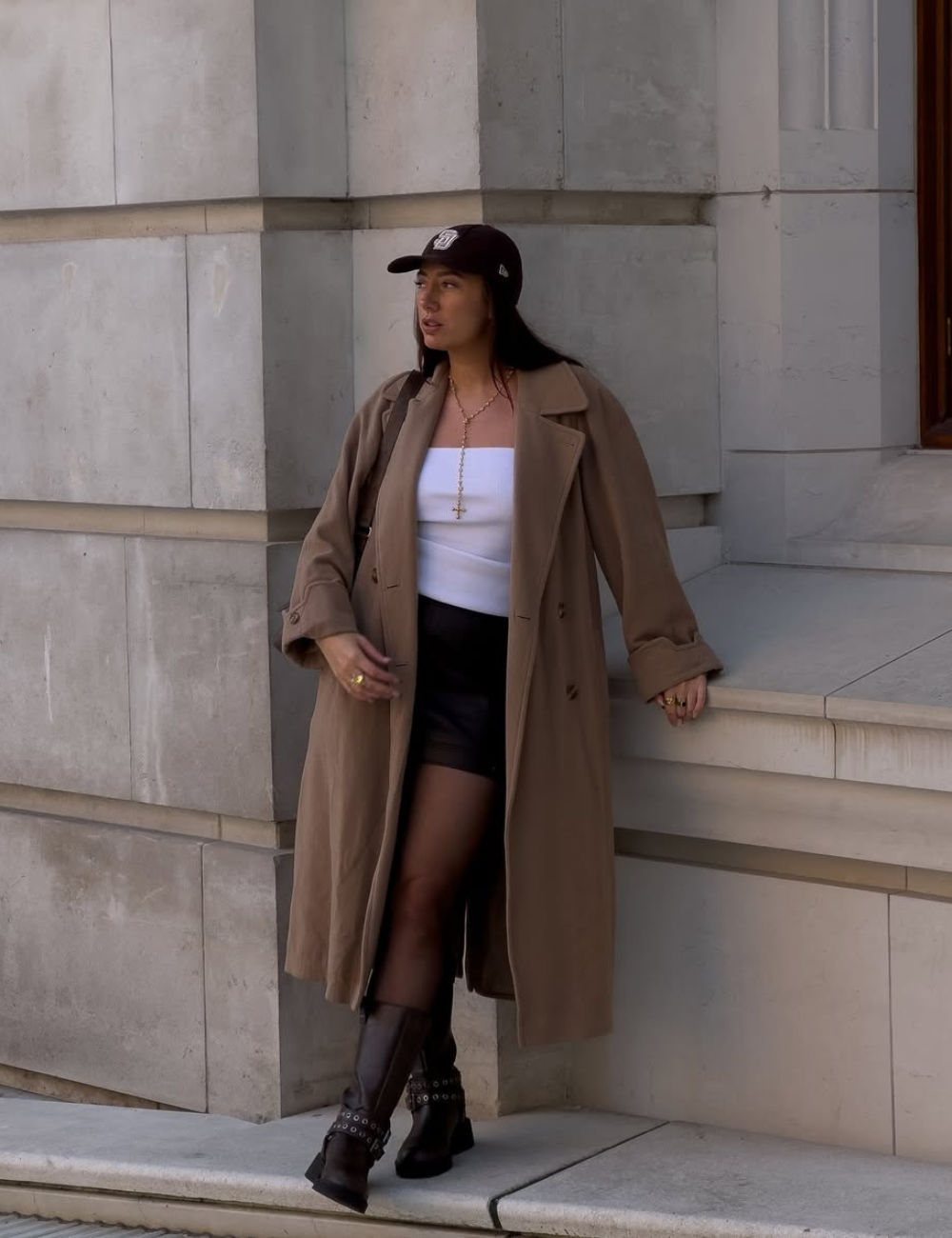 Looks com meia-calça: modelo posa com trench marrom, top branco, minissaia preta e bota robusta em cenário urbano sóbrio.