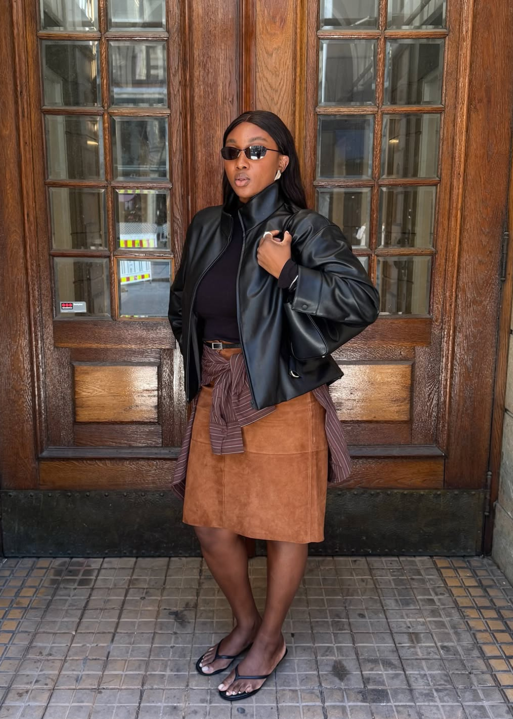 Modelo posa confiante com look outonal que combina saia lápis caramelo em camurça, jaqueta preta de couro e blusa gola alta.