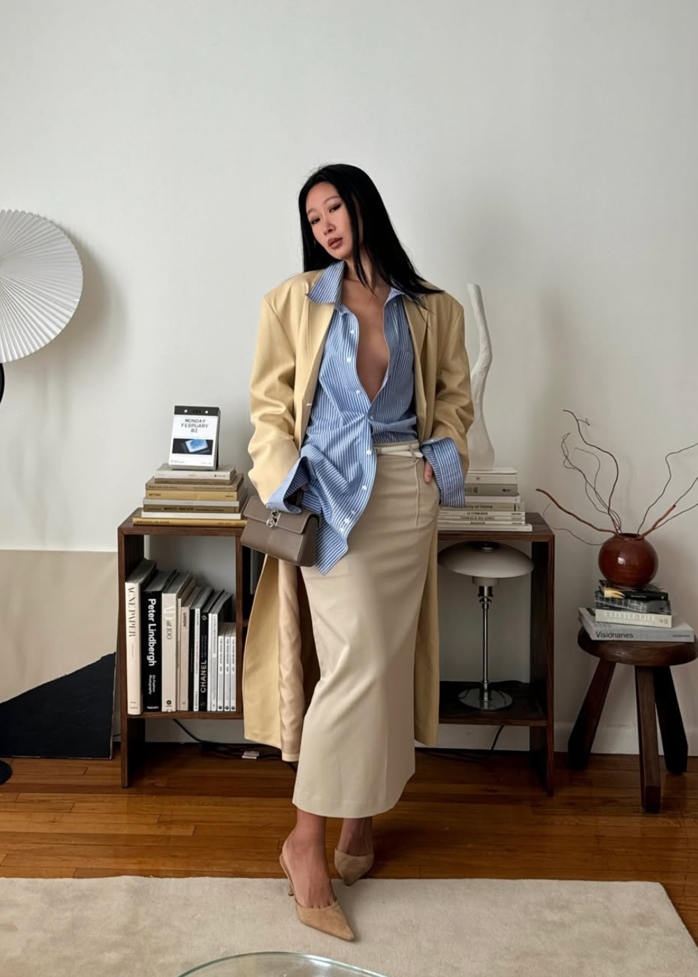 Modelo posa com look de saia lápis bege de couro, camisa azul listrada oversized e sobretudo amarelo claro.