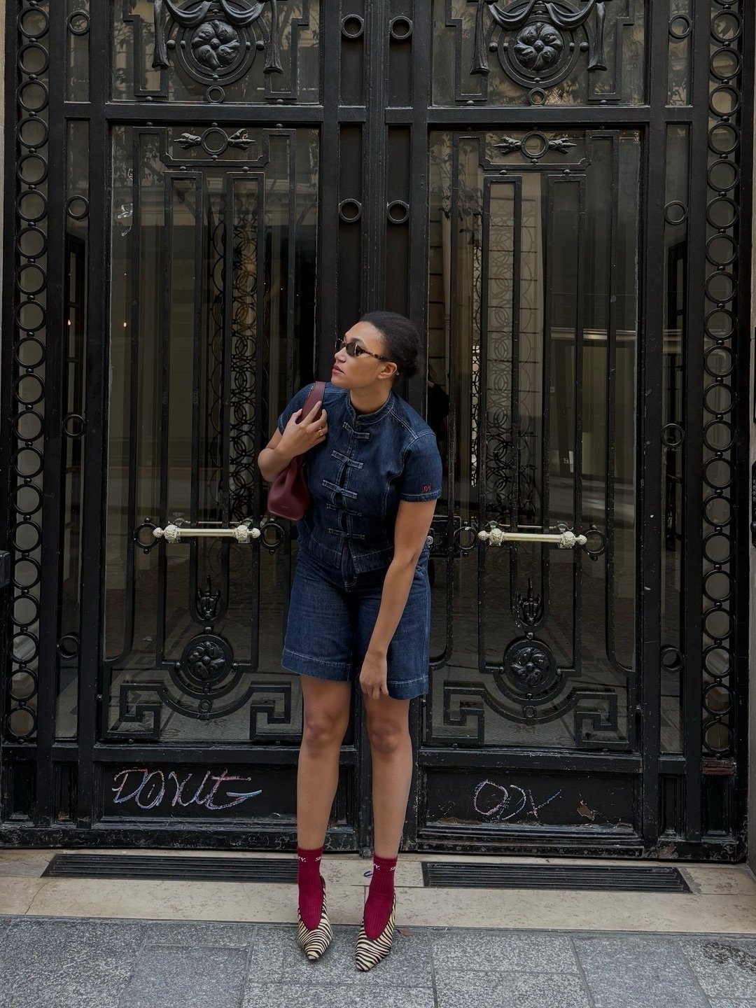 Looks criativos: modelo posa de macaquinho jeans escuro, bolsa vinho e scarpin zebra diante de portão preto imponente.