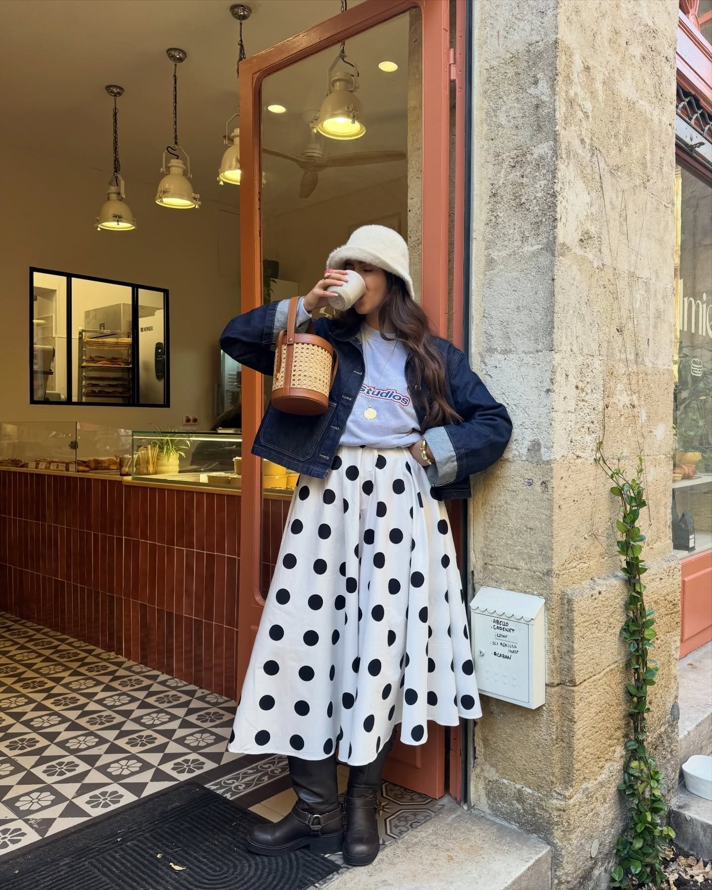 Looks criativos: modelo posa com jaqueta jeans, saia poá e bucket off-white na porta de café acolhedor.