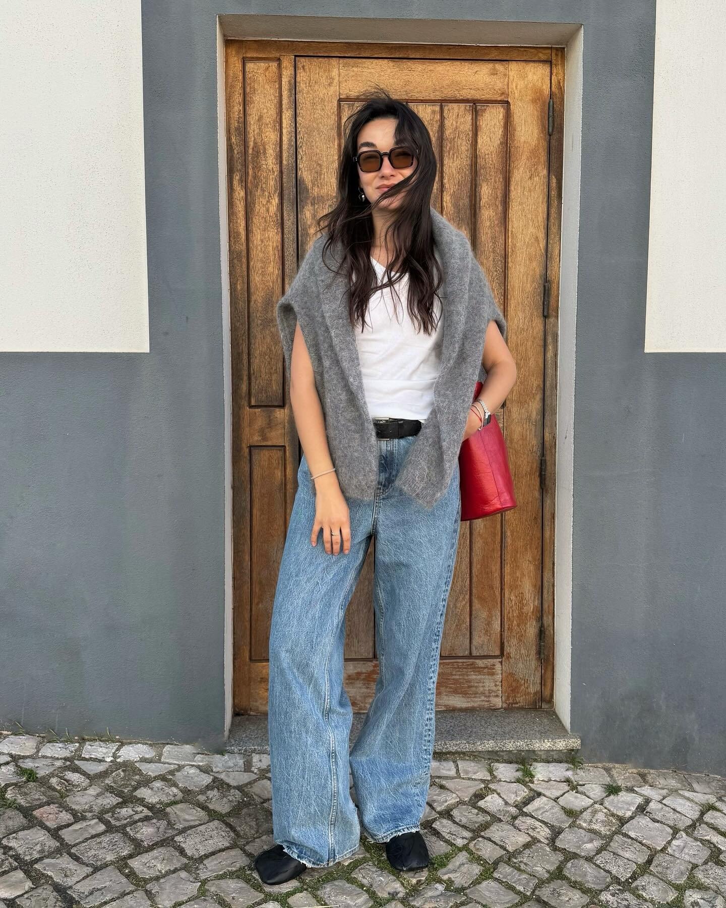 Look para faculdade com jeans largo, camiseta branca, suéter cinza sobre os ombros, bolsa vermelha e óculos escuros