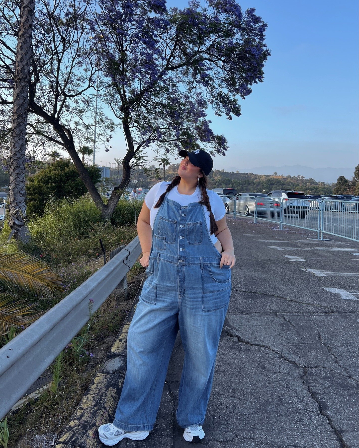 Looks para faculdade com jardineira jeans ampla, camiseta branca justa, tênis esportivo branco e boné preto