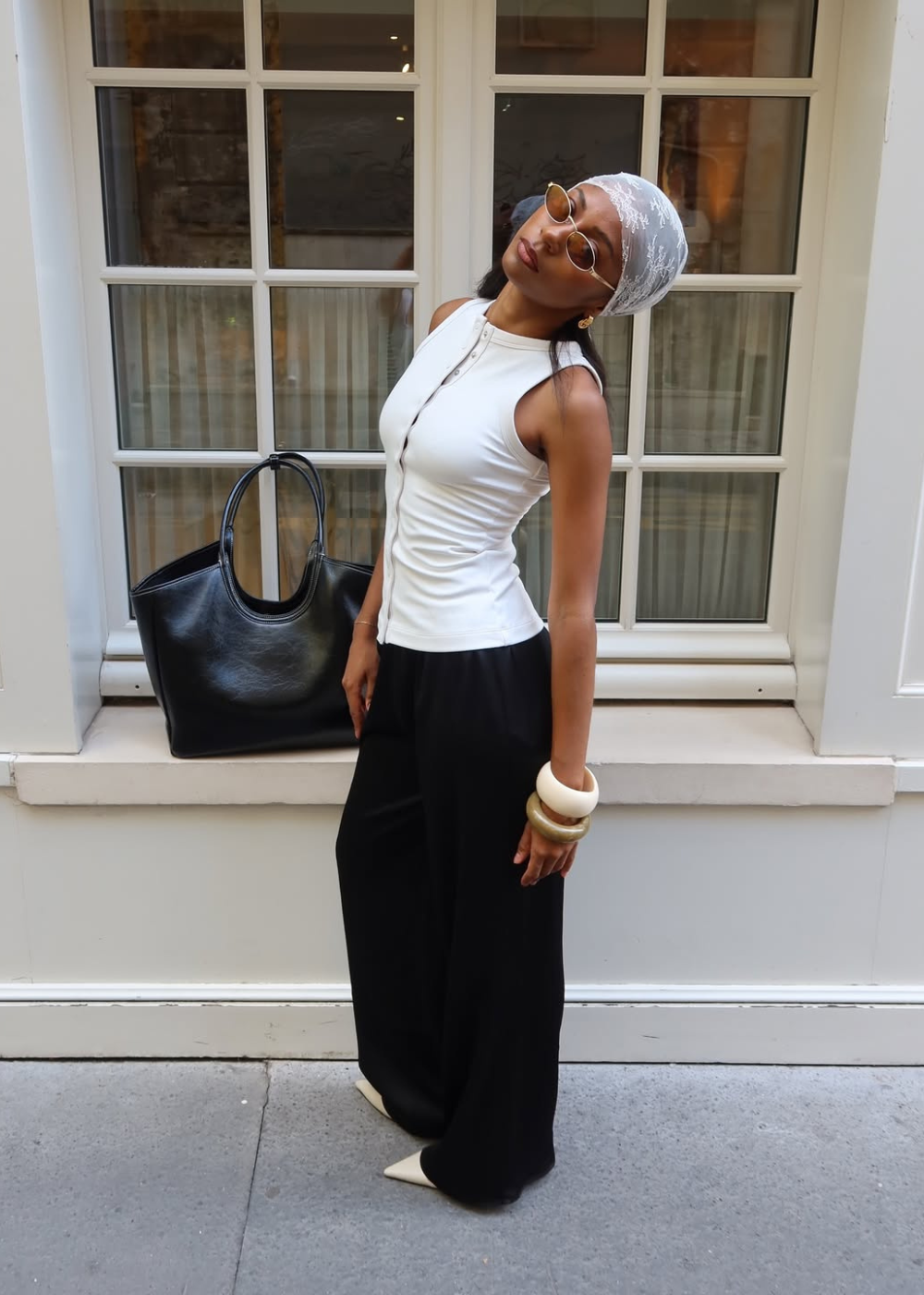 Modelo posa com postura relaxada usando regata branca justa de botões, calça preta ampla, lenço branco de renda e acessórios.