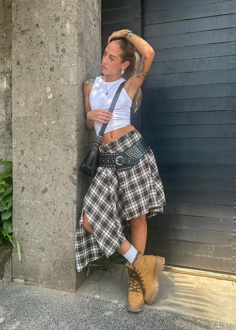 Looks para o Lollapalooza 2026 com top branco justo, saia assimétrica xadrez marrom, cinto preto com tachas e coturno.