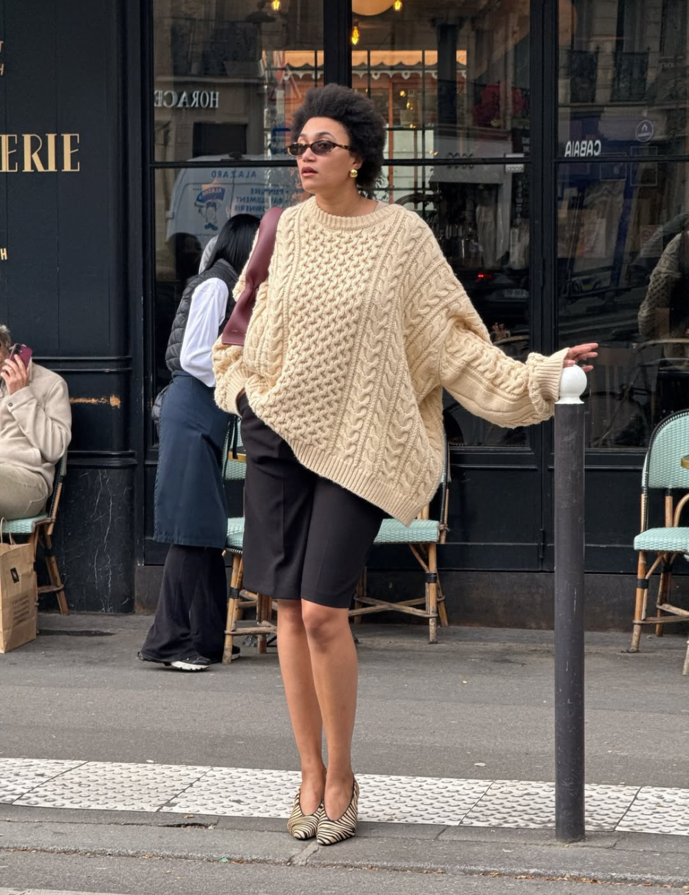 Looks de meia-estação: modelo posa com tricô oversized bege, bermuda preta e scarpin animal print na calçada urbana.