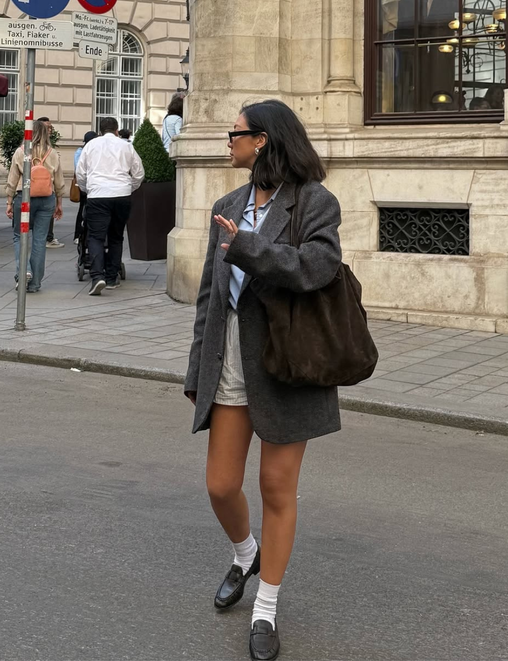 Looks de meia-estação com blazer cinza oversized, camisa azul, short claro e mocassins; modelo posa com elegância urbana.