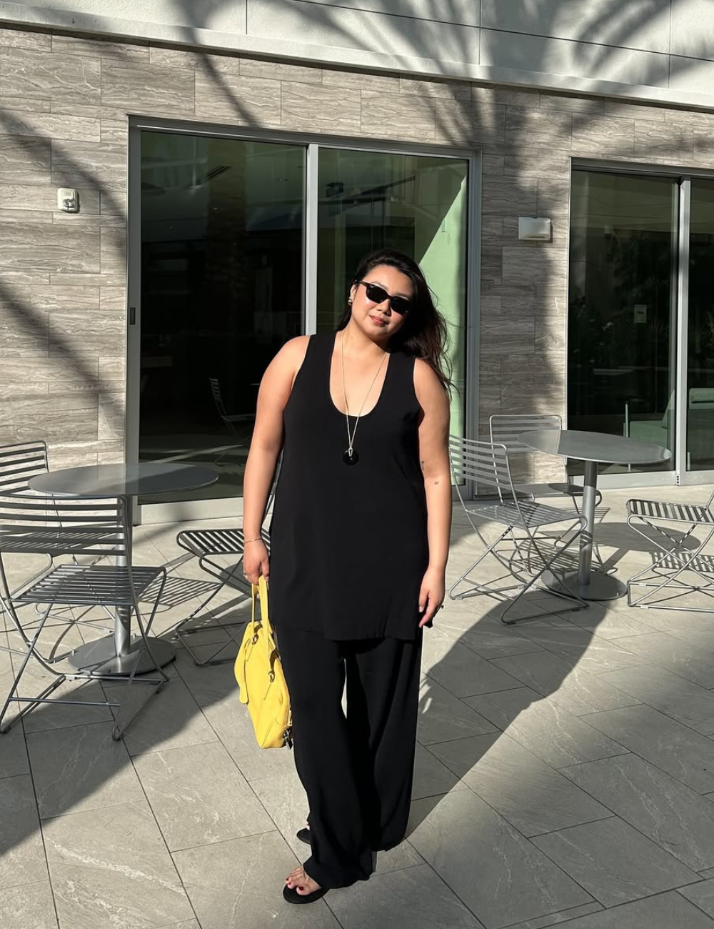 Looks de meia-estação com modelo posa de macacão preto fluido, óculos escuros e bolsa amarela em varanda ensolarada.