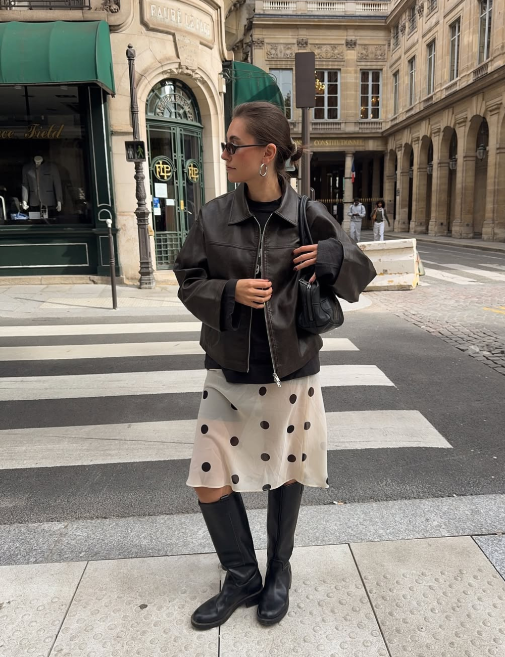 Looks de meia-estação com modelo posa de jaqueta marrom, saia poá e botas pretas em rua clássica, ar elegante.