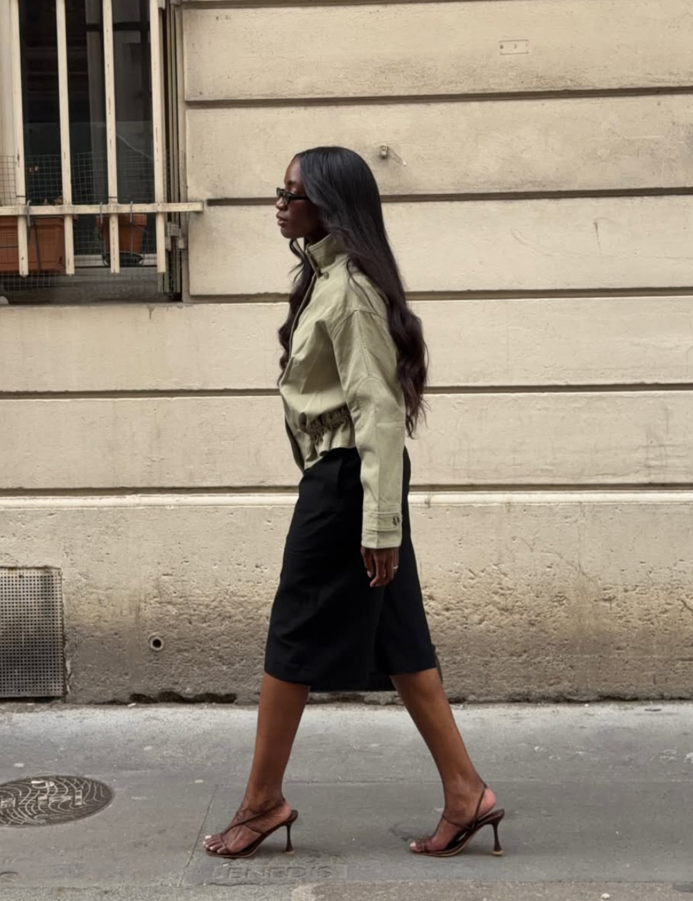 Looks de meia-estação com modelo caminha em jaqueta verde cropped, saia preta midi e sandálias, em clima urbano elegante.