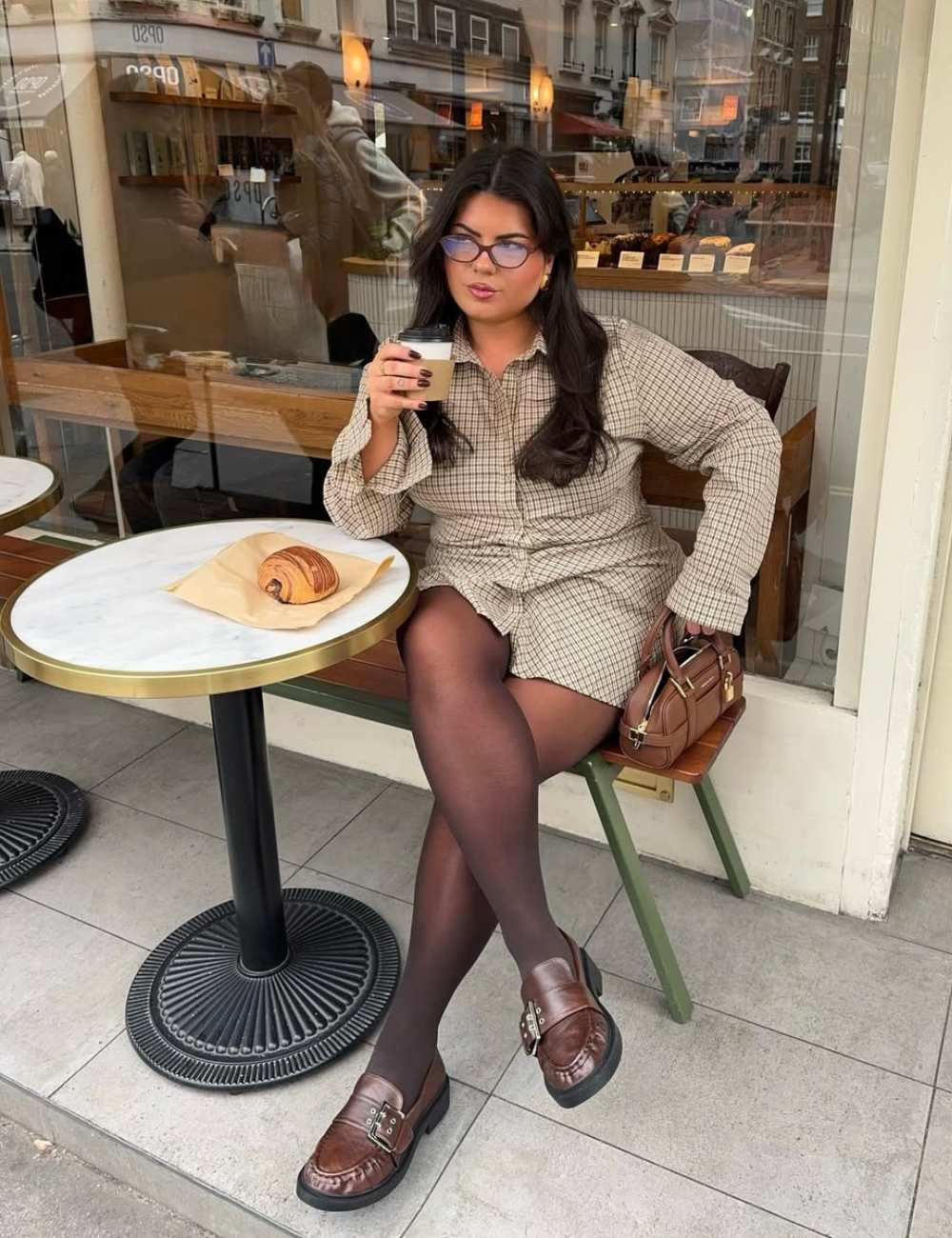 Looks de meia-estação com vestido xadrez, meia-calça e mocassim marrom; modelo posa em café com elegância casual.