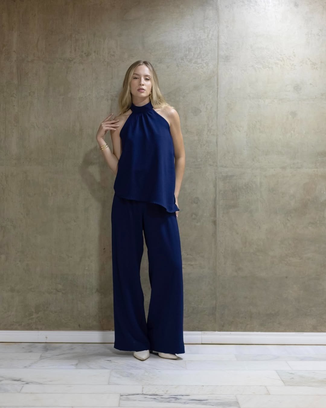 Modelo posa com look com calça pantalona preta de tecido fluido, colete ajustado e acessórios prateados, em clima elegante e