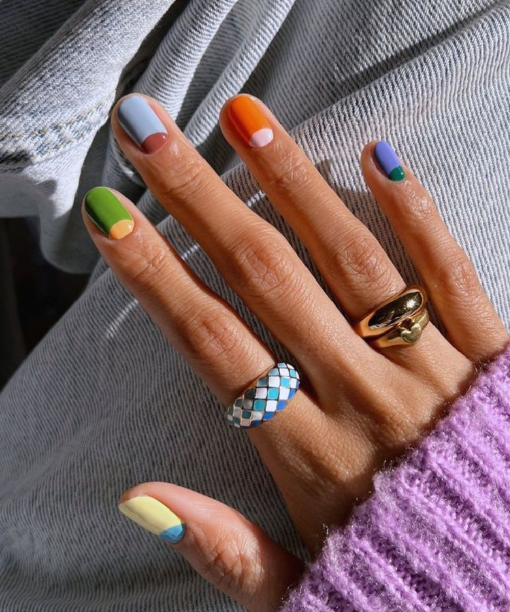 Esmaltação polêmica com design geométrico em cores vibrantes e anéis dourado e esmaltado, sobre suéter lilás e jeans claro.