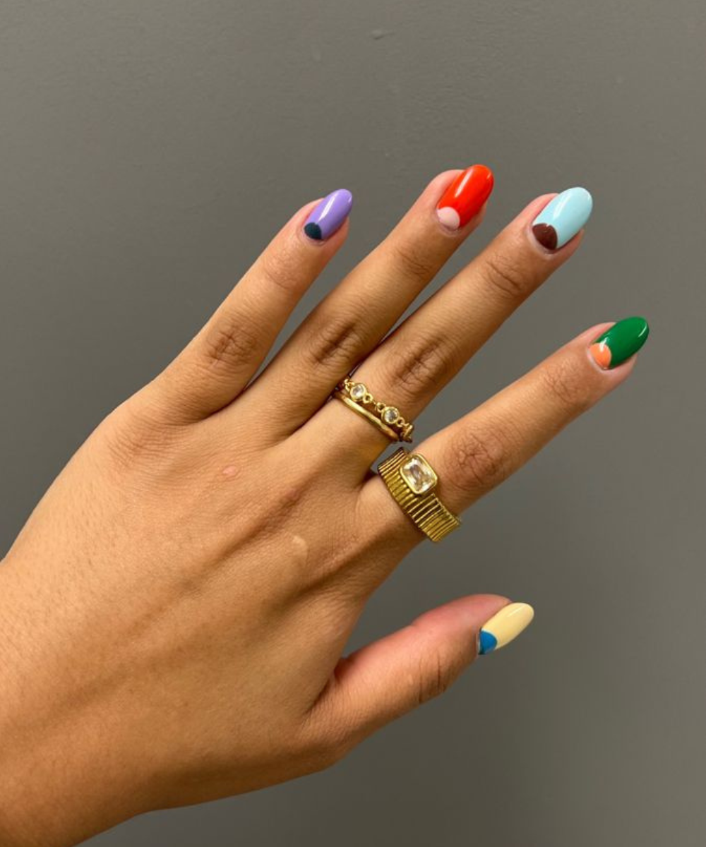 Mão com esmaltação polêmica em tons vibrantes e formas geométricas, adornada por anéis dourados que realçam estilo moderno e