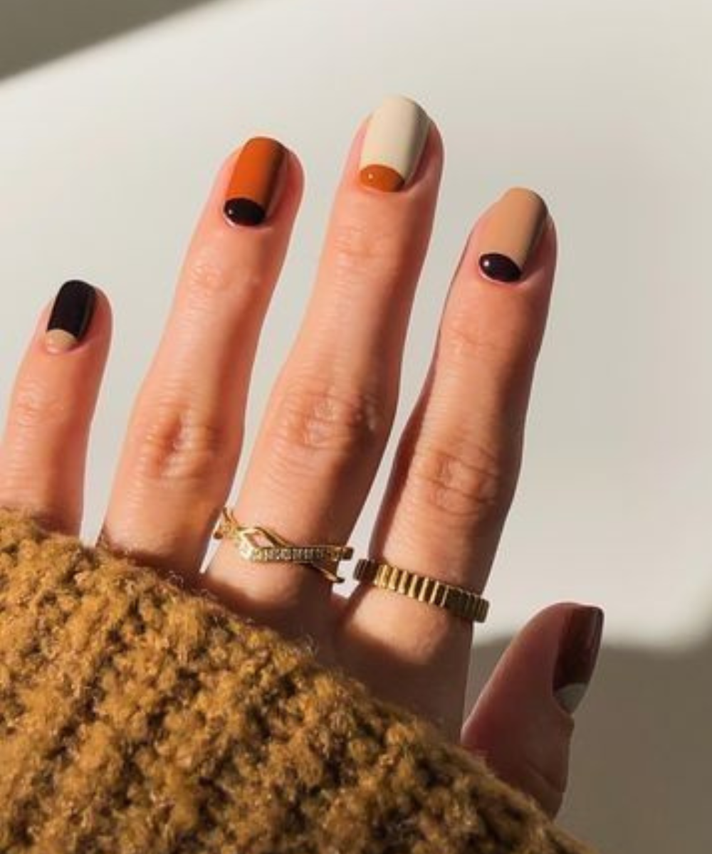 Esmaltação polêmica com design geométrico em tons terrosos e preto, unhas curtas brilhosas e anéis dourados delicados.